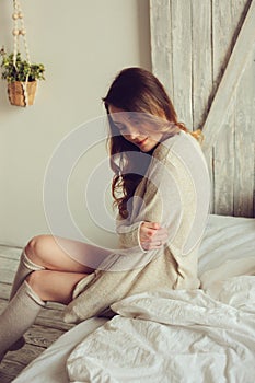 Young woman in knitted cardigan and warm socks wake up in the morning in cozy scandinavian bedroom and sitting on bed with white b