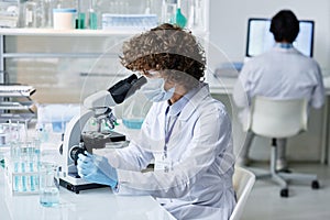 Young woman in labcoat, protective mask and gloves looking in microscope