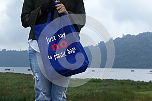 Young woman on the lake holding a blue women foldable shopping bag reusable eco with text message - Just say no to plastic bags.