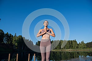 Young woman by the lake practicing yoga moves on wooden platform. Pretty young woman exercising in nature, healthy