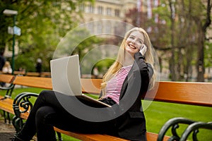 Young woman with laptop on the bench using mobile phone amd sitting in the park