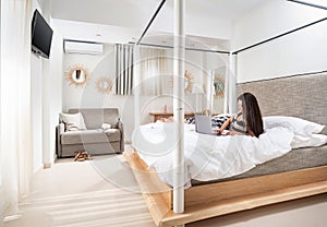Young woman with laptop laying on white bed in modern design bedroom interior with light gray and natural wood furniture