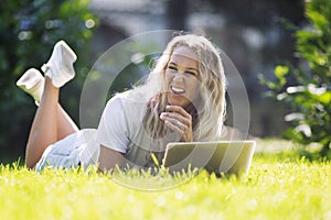 Young woman with laptop lies on the grass in a summer park. Laughing blonde with long hair in a white tank top and shorts. Sunny