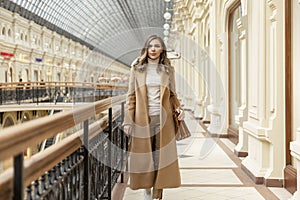 Young woman in a large light shopping mall. Beautiful smiling blonde in a beige coat. Full height
