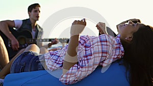 Young woman laying on easychair and listening to a man playing guitar among group of friends sitting on the beach and