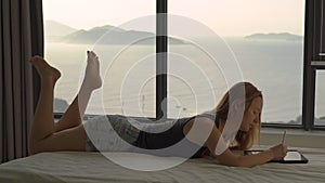 A young woman lays on a bed and uses a tablet behind her there is a window with a panoramic view on a coast and a rising
