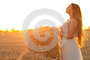 Young woman lean on haystack walking in summer evening, beautiful romantic girl outdoors in field at sunset