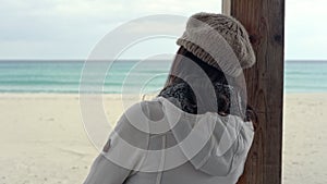 Young woman leaning against a wooden pole in the sunroom of a closed bar by the sea in winter with a stormy sky, looks at the