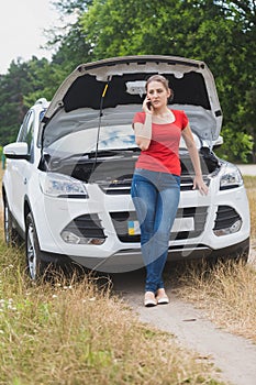 Young woman leaning on broken car and talking by phone