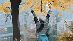 Young woman in leather jacked jumps from excitement under the autumn colored tree in the city park