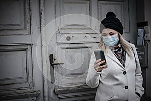 Young woman leaving house in winter, reading messages and wearing face mask due to covid-19 pandemic