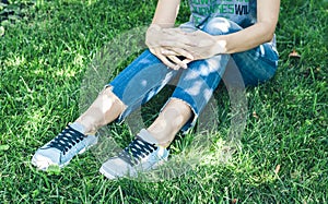 Young woman legs in sport shoes sneakers of blue suede, sitting on the grass lawn in park