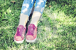 Young woman legs in sport shoes sneakers of pink suede, sitting on the grass lawn in park