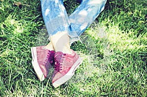 Young woman legs in sport shoes sneakers of pink suede, sitting on the grass lawn in park