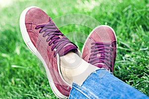 Young woman legs in sport shoes sneakers of pink suede, sitting on the grass lawn in park