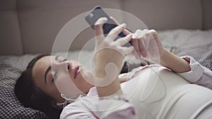 Young woman lies on a bed with a smartphone in hands, earphones plugged-in, girl listens to the music or podcast.