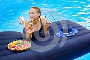 Young woman lies on a blue air mattress and relaxes in the pool and drinks a cool cocktail