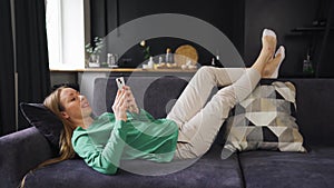 Young woman lies on the couch at home with a phone, resting, shopping, chatting, playing games