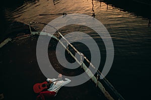 A young woman lies with guitar on an old abandoned ship against water background