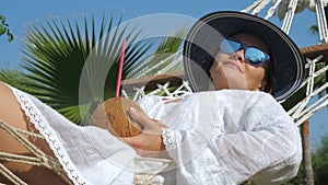 Young woman lies in a hammock with coconut water cocktail.