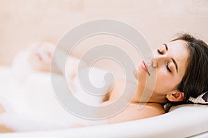 Young woman lies in white bath of foam with her eyes closed. Spa procedures, cleansing, relaxation, self-care.