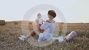 A young woman in a light dress is sitting on the grass in the field and plays with a lovely baby. Happy mother with son