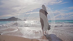 A young woman in a light transparent summer dress walks along the beach towards the sea. Sunny and windy weather. Summer beach