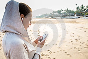 Young woman is listening to the music on the phone before jogging on the beach at the sunrise