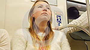 Young woman listening to music in the subway car. Media. Girl wearing white sweater with pink headphones sitting in
