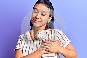 Young woman listening to music using headphones smiling with hands on chest, eyes closed with grateful gesture on face