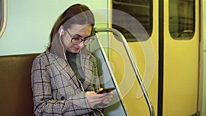 A young woman listens to music on headphones with a phone in her hands in a subway train. The girl is in correspondence
