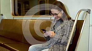 A young woman listens to music on headphones with a phone in her hands in a subway train. The girl is in correspondence