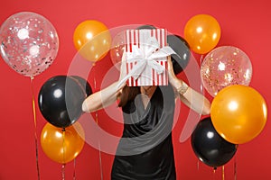 Young woman in little black dress celebrating, covering face and hiding behind red box with gift, present on bright red