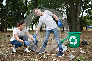 A young woman and a little boy picking up the trash and putting it in a black garbage bag on a blurred park background.