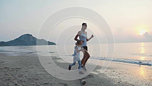 Young woman with little daughter jogging together on the beach at sunrise