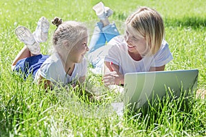 Young woman with a little daughter lie in the park with a laptop and laugh. Blogging, online training and distance learning