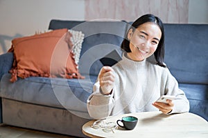 Young woman in living room, showing her credit card and using smartphone to pay, order online or shopping on application