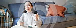 Young woman in living room, showing her credit card and using smartphone to pay, order online or shopping on application