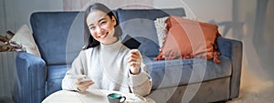 Young woman in living room, showing her credit card and using smartphone to pay, order online or shopping on application