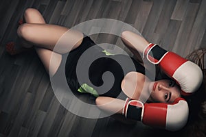 Young woman with long blonde hair and closed eyes, dressed in fitness clothing and wearing red boxing gloves, lying on dark floor