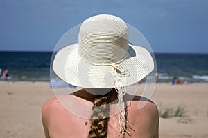 A young woman with a long braid in a white hat sitting on the beach looking at the sea