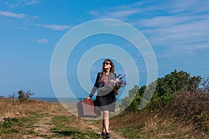 Woman with long brown hair in denim jacket, black skirt, vintage suitcase, flowers bouquet off-road. Lifestyle photo