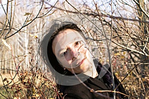 A young woman with long brown hair wearing a black coat stands in the middle of some bushes looking down, lost in thought.