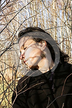 A young woman with long brunette hair is standing near the bushes in a park.