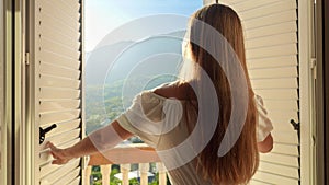 Young woman in a long dress gracefully opens balcony doors, steps onto the terrace, and embraces the mesmerizing