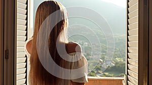 Young woman in a long dress opens her balcony doors and ventures onto the terrace, where she gracefully welcomes the