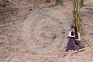 Young woman in long dress reads a book sitting under a tree