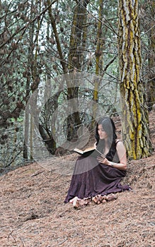 Young woman in long dress sits under a tree with book