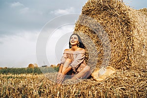 A young woman with long hair and in a dress sits near a hay bale. Woman posing smiling and looking at camera
