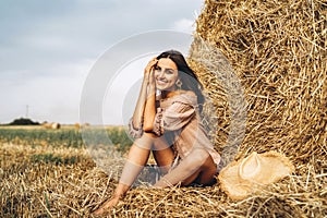 A young woman with long hair and in a dress sits near a hay bale. Woman posing smiling and looking at camera